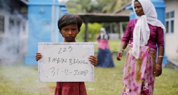 A Rohingya migrant mother stands nearby as her child holds a placard while posing for photographs for immigration purposes.