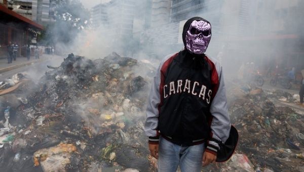 A masked opposition protester stands in front of a burning pile of garbage built to block the street.