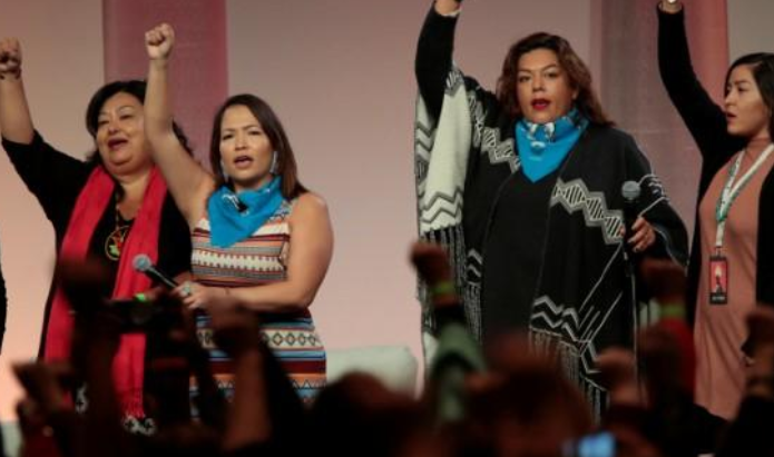 A group of Indigenous women raise their fists as they sing during the opening session of the three-day convention at Cobo Center in Detroit, Michigan.