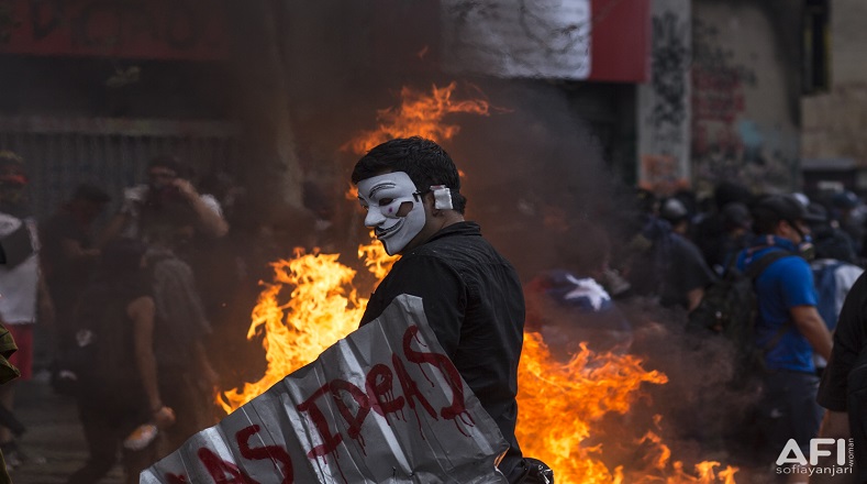 Manifestante con máscara del personaje V de vendetta, y un escudo artesanal con las palabras "Las ideas", se acerca a una barricada mientras, en la Plaza Italia, carabineros y manifestantes se enfrentan (Santiago de Chile, 8 de noviembre de 2019).