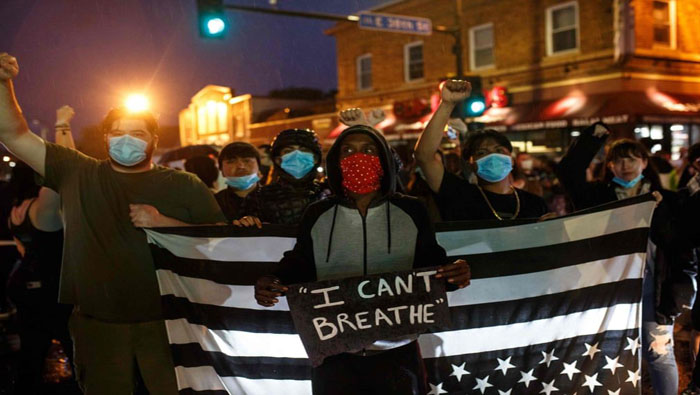 Un manifestante afroamericano sostiene un cartel con el lema inmortalizado por Floyd "No puedo respirar". Detrás del sujeto se avista la bandera de EE.UU. con las estrellas hacia abajo, y los colores blanco y negro, lo cual refleja el conflicto racial estadounidense. 